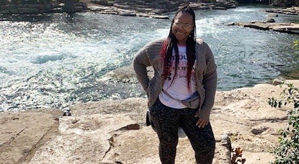 Michelene Holmes standing outside with a lake behind her. She has one hand on her back and one on her leg. She is a Black woman with long black hair.