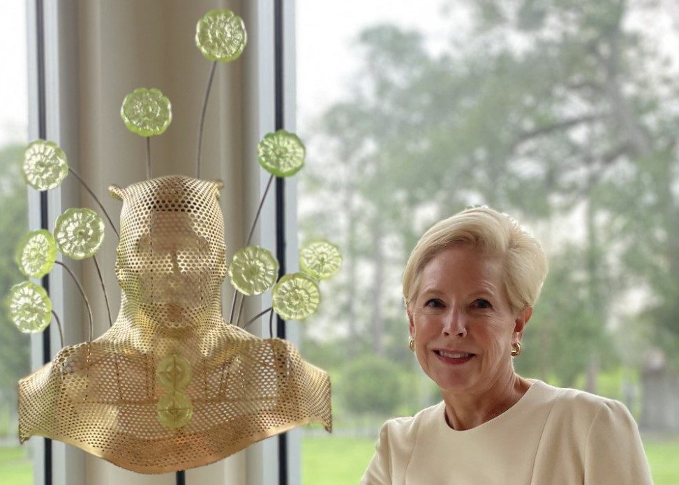 Roberta Burns stands next to a gold sculpture of her radiation mask adorned with green flowers made of uranium glass